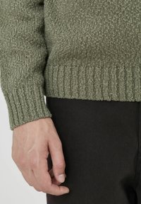 Pull en maille verte texturé avec des poignets et un ourlet côtelés, présentant un design en tricot lâche. La main s'approche d'un pantalon sombre.