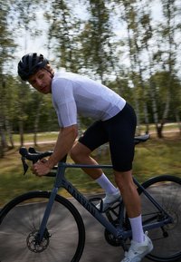 Szary rower Canyon jest jeźdzony przez rowerzystę w białej koszulce z krótkim rękawem i czarnych szortach, z białymi skarpetkami i czarnym kaskiem.