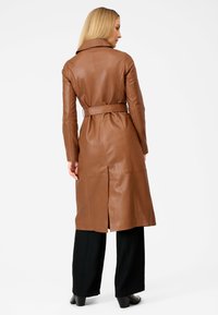Brown leather trench coat with a belted waist, long sleeves, and a collar. Features a center back vent and a smooth texture. Black pants visible below.