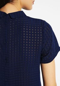 Marinblå kortärmad blus med ett rutigt genomskinligt tyg, klassisk krage och knappstängning baktill.