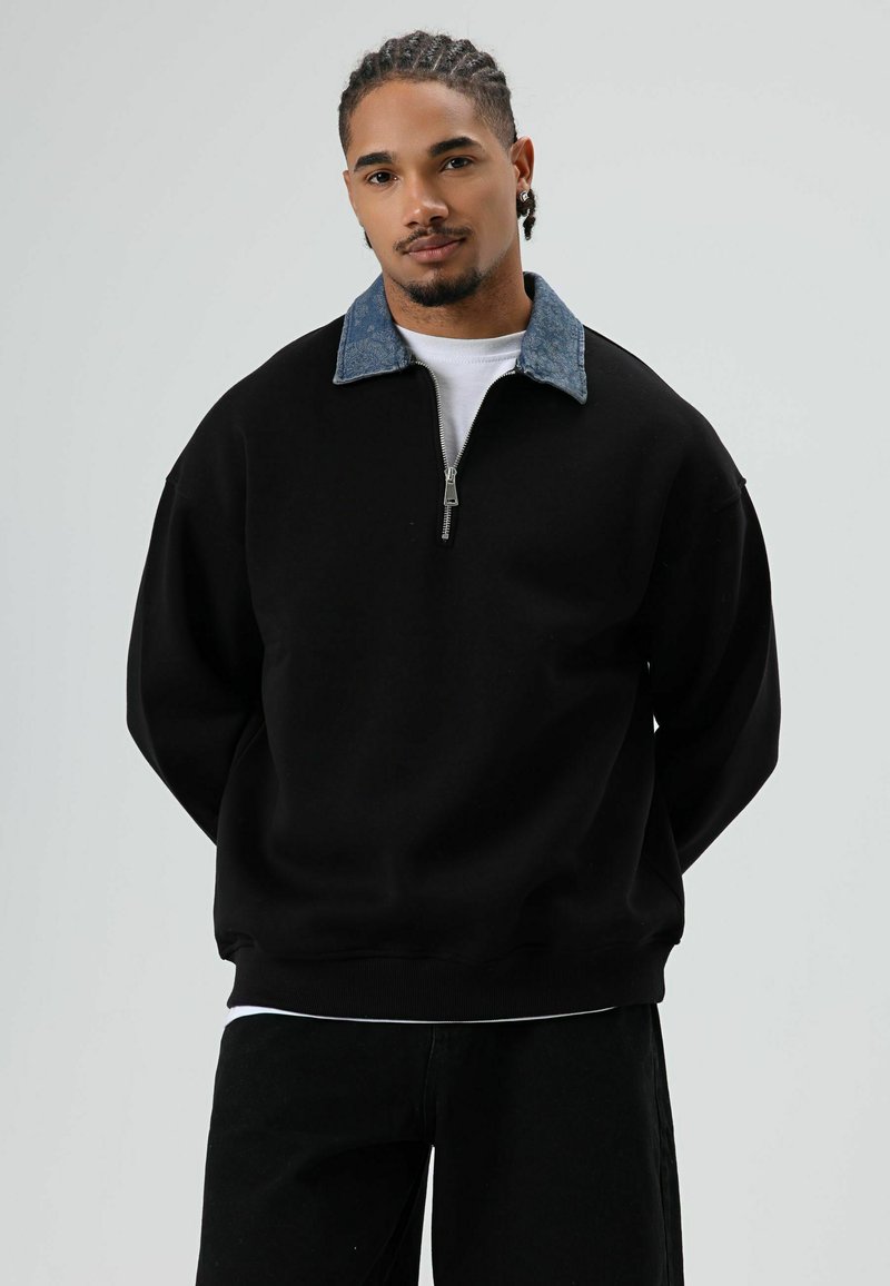 Zwarte sweatshirt met een kwartrits en een denim kraag. Heeft een relaxte pasvorm en geribbelde manchetten, met contrasterende donkere en lichte texturen.