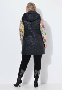Kvinna i svart quiltad väst med huva, färggad långärmad topp, svarta leggings med strassrosetter och svarta högklackade skor, vänd bort.