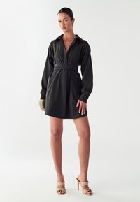 Mustaga kleit koos krae, pikkade varrukate ja vöödiga taljehoga. Sile kangas ja nööpidega esiosa. Kantud beeži kontsaga sandaalidega.