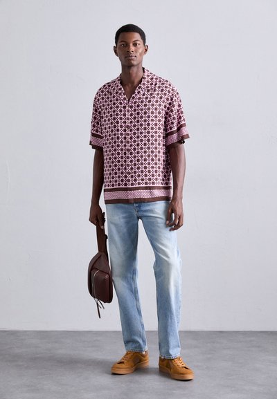 Chemise à manches courtes marron à motifs avec des accents roses, jean bleu clair et baskets marron, tenant un sac marron, debout sur une surface grise.