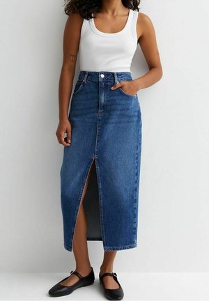 Frau trägt ein weißes Tanktop, einen hoch taillierten blauen Jeans-Midirock mit vorderem Schlitz und schwarze Mary Jane Ballerinas, steht vor einer weißen Wand.