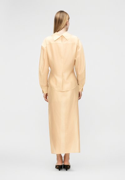 Γυναίκα με ίσια ξανθά μαλλιά, φορώντας μια μπεζ μπλούζα μακρυμάνικη και μακρύ φόρεμα στο ίδιο χρώμα, όρθια με την πλάτη στραμμένη, φαίνονται μαύρες γόβες.