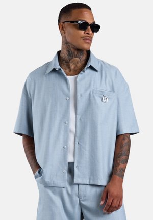 Mann mit Hals- und Arm-Tattoos, trägt schwarze Sonnenbrille, hellblaues kurzärmeliges Hemd mit Knöpfen und passende Hose, steht mit den Händen in den Taschen.