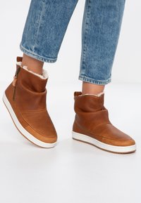 Botas de tobillo de cuero marrón con forro de piel sintética, que presentan un diseño plegable de moda y una suela de goma blanca con agarre texturizado.