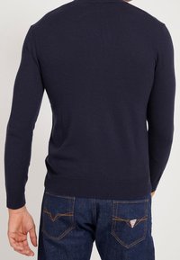 Uomo che indossa un maglione blu notte e jeans blu con tasche posteriori cucite, rivolto di spalle contro uno sfondo chiaro e semplice.