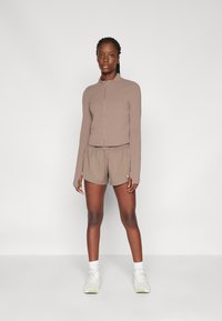 Hnědý zip-up top s dlouhými rukávy a odpovídající šortky vyrobené z lehkého materiálu. Šortky mají elastický pas a obsahují malý detail loga.