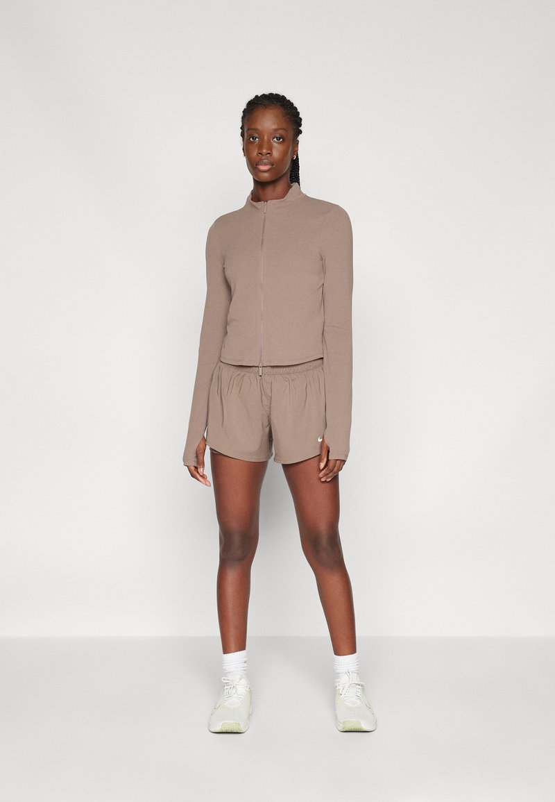 Top a maniche lunghe con zip di colore marrone e pantaloni corti abbinati, realizzati in tessuto leggero. I pantaloni corti hanno una vita elastica e presentano un piccolo dettaglio con logo.