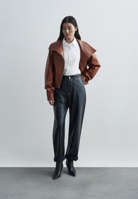 Veste en cuir marron avec large col, chemise blanche boutonnée et pantalon en cuir noir taille haute. Des talons pointus noirs complètent la tenue.