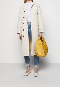 Trench-coat beige clair avec de gros boutons marron, chemise blanche, jean bleu déchiré et sac en cuir jaune. Des chaussures plates blanches complètent le look.