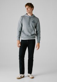 Felpa grigia in pile con tasca frontale, dettaglio del logo nero e cordino regolabile, abbinata a jeans scuri e sneaker con collo bianco.