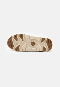 Schuhsohle mit einem strukturierten Design. Hauptfarbe ist Creme mit braunen Gummielementen. Verfügt über tiefe Profilrillen und ein kreisförmiges Logo in der Mitte.