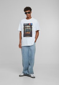 Hvid bomulds t-shirt med et trykt metro-scene. Løst siddende lyseblå denimbukser og sorte solbriller, båret med grå og hvide sneakers.