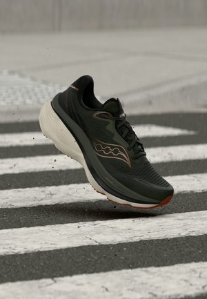 Chaussure de course noire avec une semelle blanche épaisse en l'air au-dessus d'un passage piéton en ville, avec de petits débris soulevés en dessous.