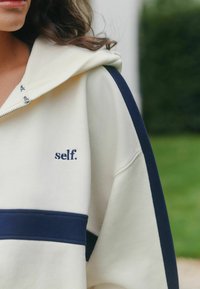 Krémová mikina s kapucňou s námorníckymi detailmi a predným zipsom. Na ľavej hornej strane je vyšitý nápis "self." v námorníckej modrej farbe. Mäkká textúra.