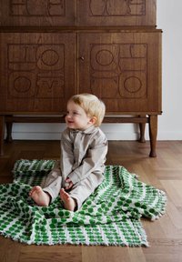 Barn siddende på et grøn- og hvidt bladmønstrede tæppe, iført en beige knap-op outfit, med træmøbler i baggrunden.