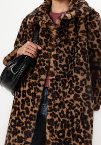 Nep-bont jas met luipaardprint en een kraag, voorzien van knopen en een losse pasvorm. Een persoon draagt een zwarte leren tas en een donkerrood shirt eronder.