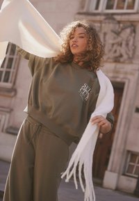 Ein olivgrüner Sweatshirt und passende Jogginghosen haben eine lockere Passform und bündchen an den Knöcheln.Ein weißer, fransiger Schal wird gehalten und hängt elegant herab.