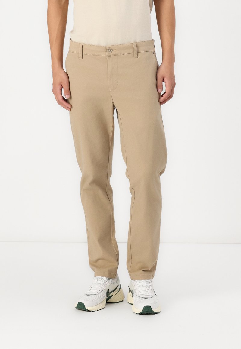 Beige chino nadrág, egyenes szabással, elöl gombos záródással és övhurkokkal. Fehér és zöld sportcipővel viselve.