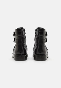 Botas de tobillo de cuero negro con un acabado brillante, que cuentan con tres correas ajustables, acentos plateados y un tacón apilado con detalles de tachuelas.