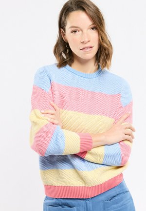 Femme aux cheveux bruns mi-longs, bras croisés, portant un pull à rayures pastel bleu, rose et jaune, debout devant un fond blanc.