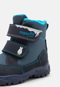 Bota de tela azul y marino con tira de Velcro, que presenta un gráfico de pingüino y costuras contrastantes, diseñada con una suela de goma texturizada.