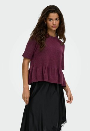 Femme aux longs cheveux ondulés portant un pull violet à manches courtes et une jupe en satin noire avec bordure en dentelle sur un fond uni.