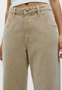 Pantalons en coton beige à taille haute, dot és d'une fermeture à bouton, de poches latérales et d'un tissu texturé. Détails de couture visibles.