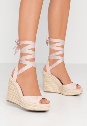 Pieds portant des sandales compensées à lacets rose clair avec des talons plateformes en jute tressé sur un fond clair.