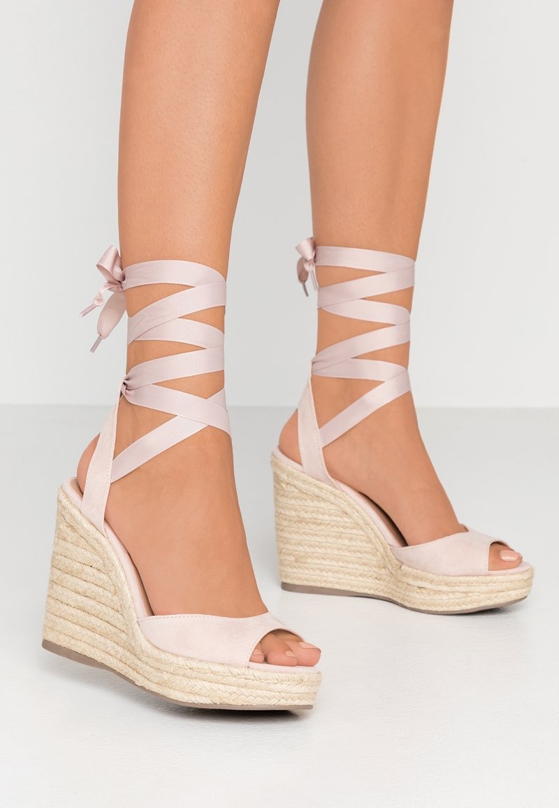 Pieds portant des sandales compensées à lacets rose clair avec des talons plateformes en jute tressé sur un fond clair.