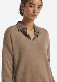 Hellbraun gestrickter Pullover mit tiefem V-Ausschnitt, der subtile strukturierte Details an den Schultern aufweist. Über einem grauen Hemd mit Kragen getragen.