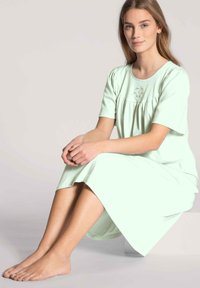 Camicia da notte verde chiaro con maniche corte, corpino plissettato e ricami floreali. Tessuto morbido, lunghezza fino al ginocchio, vestibilità rilassata. Piedi nudi visibili.