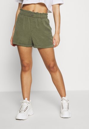 Woman wearing high-waisted olive green shorts and white chunky lace-up ankle boots, standing against a plain light background.