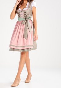 Femme portant une robe dirndl traditionnelle rose et beige avec un corsage lacé, un chemisier blanc, une ceinture verte nouée en nœud, et des talons nude.