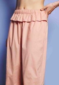 Egy személy laza, rózsaszín nadrágot visel, ráncos elasztikus derékkal, kezével kicsit meghúzza a derékrészt, egy világos lila háttér előtt.