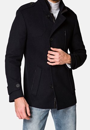 Mann trägt einen schwarzen, geknöpften Wollmantel über einem hellgrauen Rollkragenpullover und hellblauen Jeans und richtet den Mantel vorne.