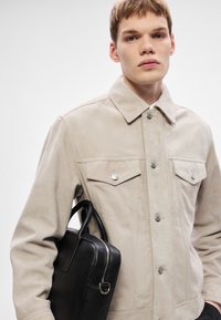 Bézs színű velúr dzseki hegyes gallérral, elülső gombokkal és két mellzsebbel. A modell egy fekete bőr aktatáskát tart, amelyen cipzár található.