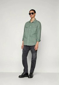 Light green button-up shirt with two chest pockets, paired with dark cargo pants and black lace-up boots. Model wearing sunglasses.