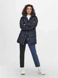 Doudoune marine avec capuche, présentant un design matelassé et des boutons-pression argentés, associée à un jean bicolore noir et bleu et des baskets blanches.