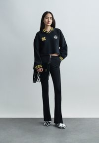 Γυναίκα που στέκεται, φοράει μαύρο κοντό πουλόβερ με κίτρινες ρίγες, μαύρο παντελόνι, ασημένια μυτερά παπούτσια και κρατάει μαύρο φάκελο με κρόσια.