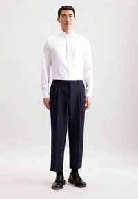 Wit cropped shirt met een opstaande kraag, gecombineerd met marineblauwe geplooide broek en zwarte leren schoenen, geplaatst tegen een lichte achtergrond.