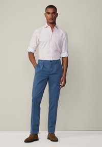 Camicia a righe rosa chiaro e bianca, maniche arrotolate; pantaloni blu con pieghe; scarpe marroni in suede; sfondo minimalista.