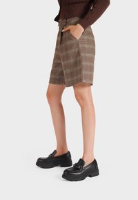 Pantaloni corti a quadri marroni con tasche laterali, abbinati a mocassini neri chunky con dettaglio in catena metallica. Tessuto liscio e vestibilità sartoriale.