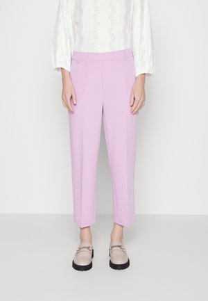 Femme portant un pantalon court violet clair, une blouse blanche texturée à manches longues et des mocassins beige avec semelles noires et détail chaîne.