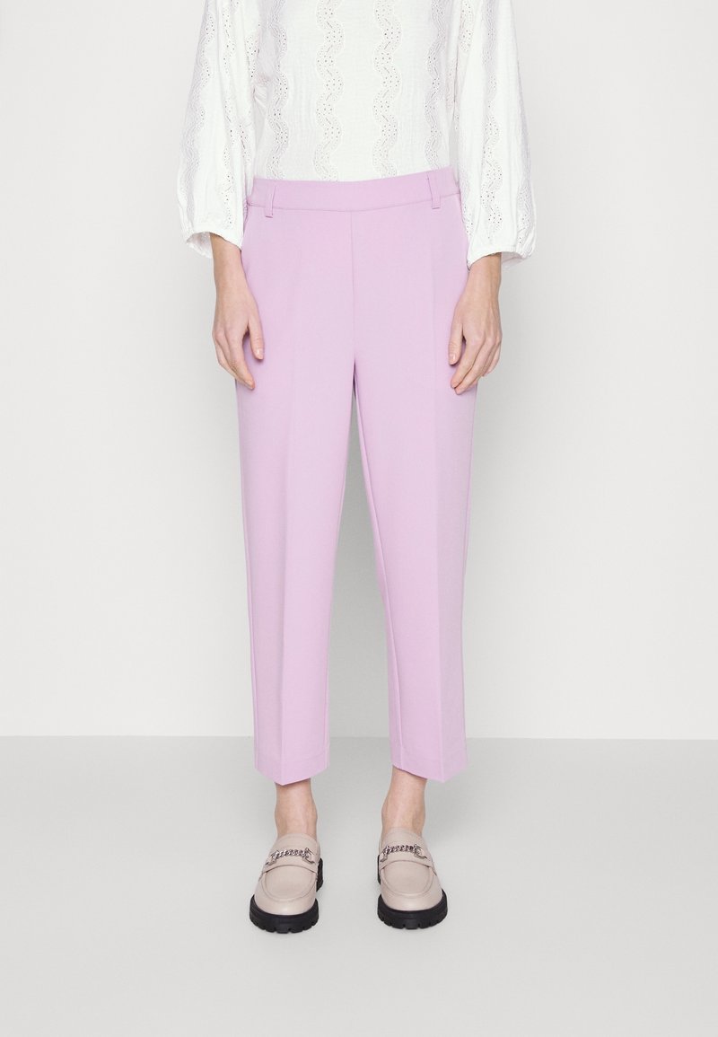 Femme portant un pantalon court violet clair, une blouse blanche texturée à manches longues et des mocassins beige avec semelles noires et détail chaîne.