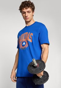 Mėlynas medvilninis marškinėliai su oranžiniu „ADIDAS“ grafiniu ženklu, trumpomis rankovėmis ir apvaliu iškirpimu. Modelis vienoje rankoje laiko juodą svorio diską.