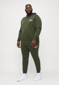 Olivgrüner Hoodie und passende Jogginghose aus weichem Stoff, mit einem Logo auf der Vorderseite und elastischen Bündchen. getragen mit weißen Turnschuhen.